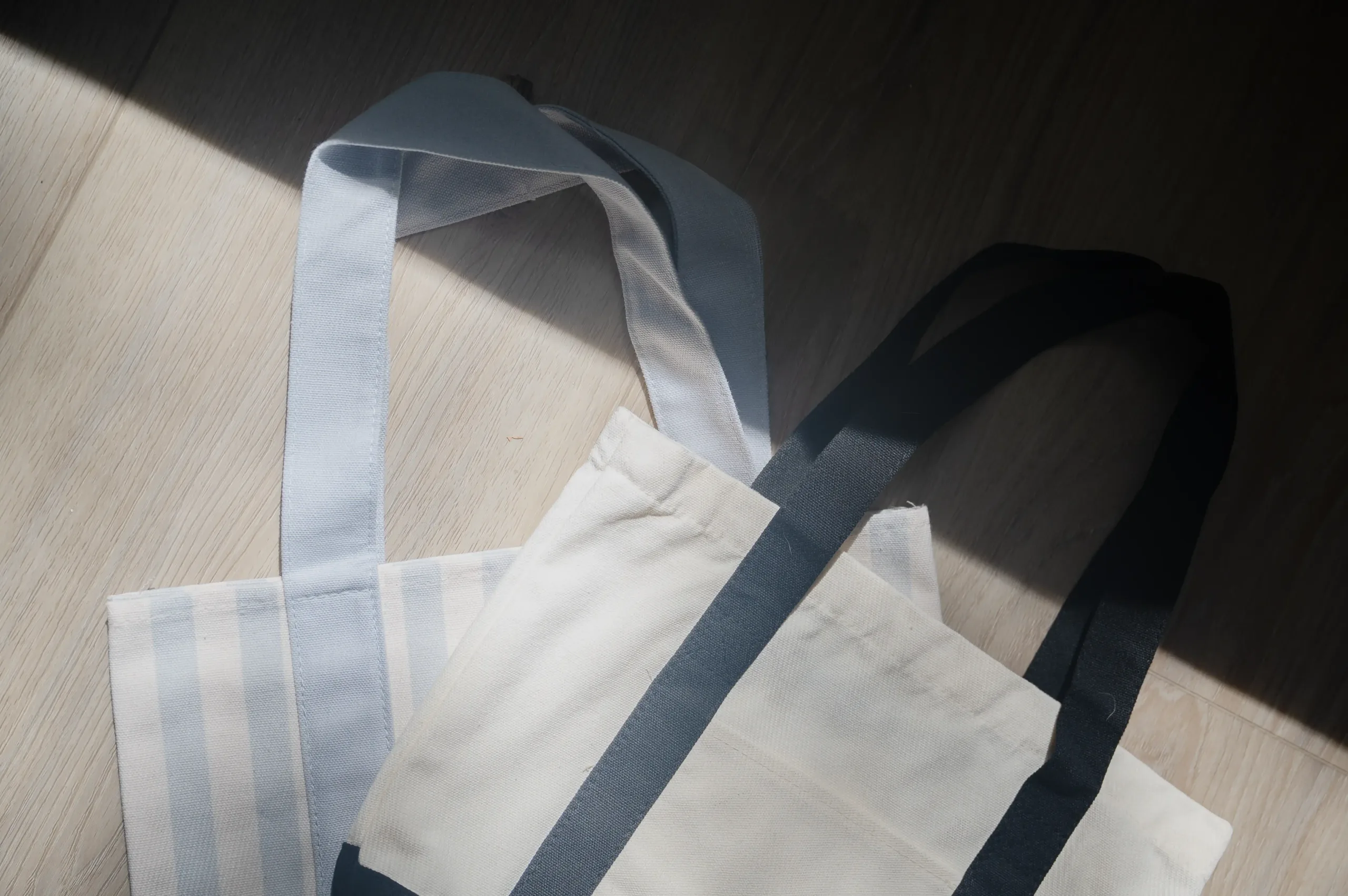 Two reusable tote bags on a wooden floor with a diagonal shadow.